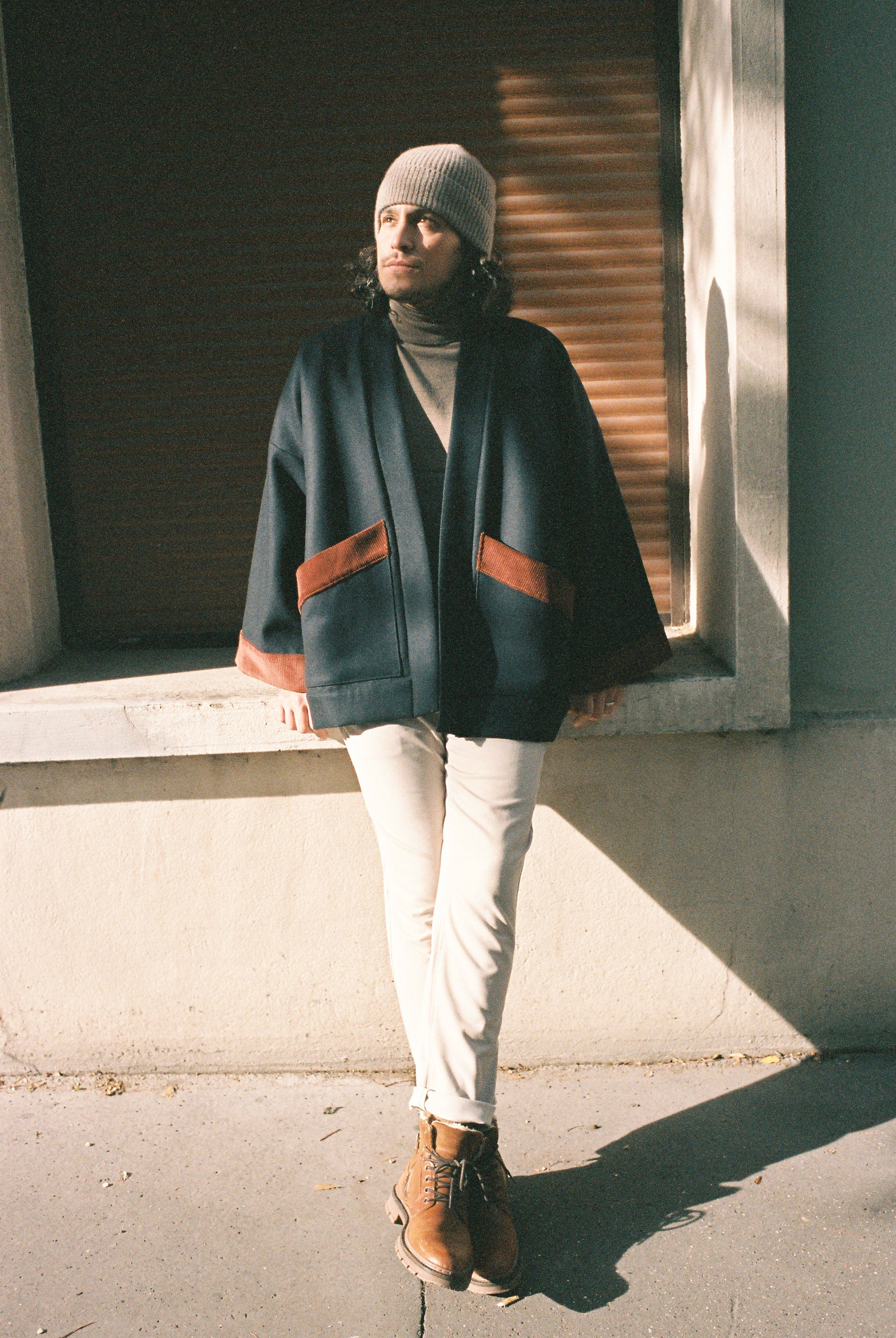 Homme portant le kimono DODAT Atelier Sarita, un kimono veste en laine caviar noire avec finitions en velours coton couleur oud, photographié en extérieur au soleil. Modèle unisexe, confectionné à la main à Paris, au style urbain et architectural.