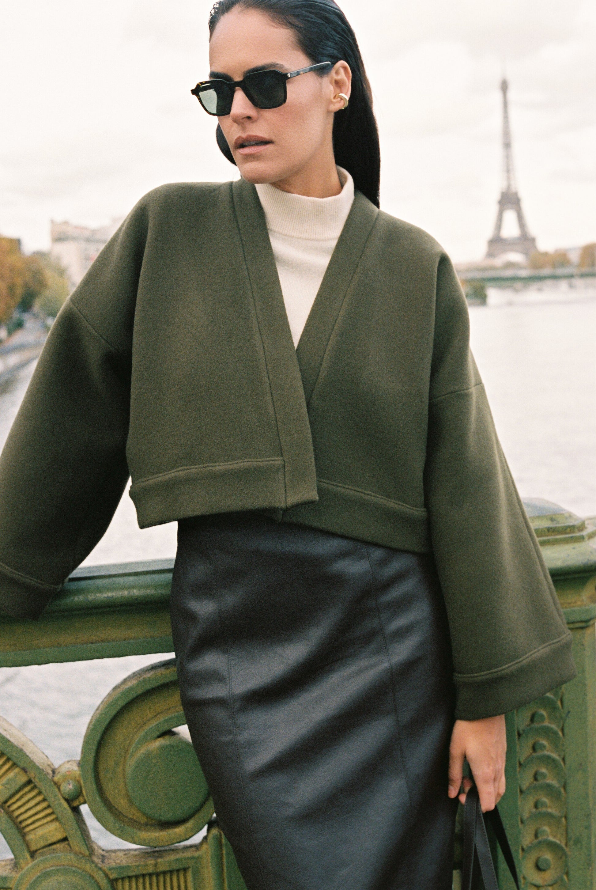 Femme portant le kimono court JOUFFROY Atelier Sarita en laine et cachemire kaki, coupe crop, avec finitions minimalistes et poches intérieures. La silhouette est photographiée sur un pont parisien avec vue sur la Tour Eiffel, mettant en valeur un kimono artisanal confectionné à la main à Paris, pensé pour l’hiver.