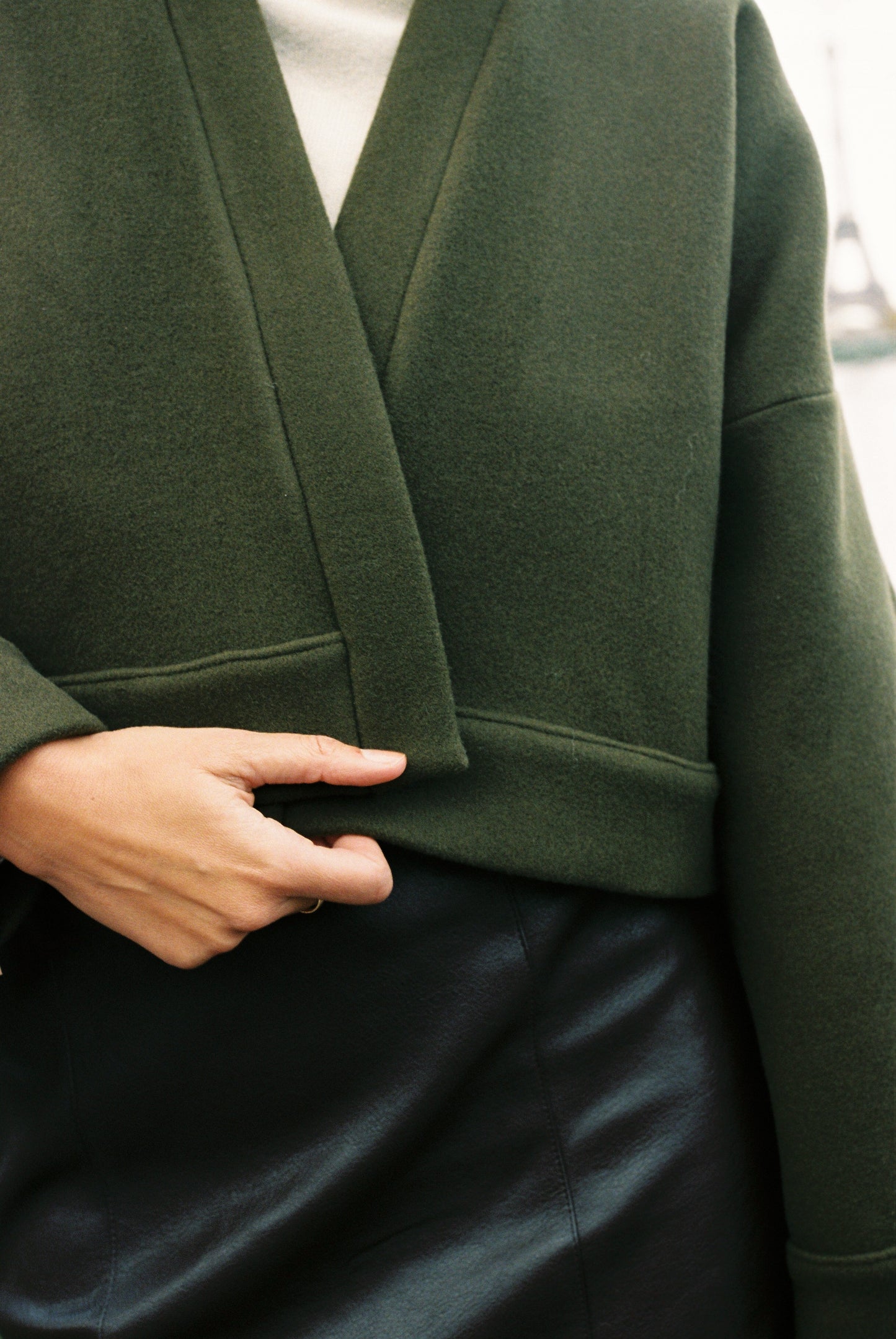 Femme portant le kimono court JOUFFROY Atelier Sarita en laine et cachemire kaki, coupe crop, avec finitions minimalistes et poches intérieures. La silhouette est photographiée sur un pont parisien avec vue sur la Tour Eiffel, mettant en valeur un kimono artisanal confectionné à la main à Paris, pensé pour l’hiver.