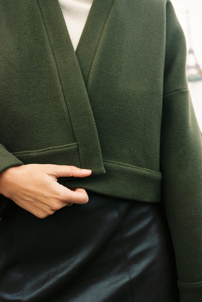 Femme portant le kimono court JOUFFROY Atelier Sarita en laine et cachemire kaki, coupe crop, avec finitions minimalistes et poches intérieures. La silhouette est photographiée sur un pont parisien avec vue sur la Tour Eiffel, mettant en valeur un kimono artisanal confectionné à la main à Paris, pensé pour l’hiver.