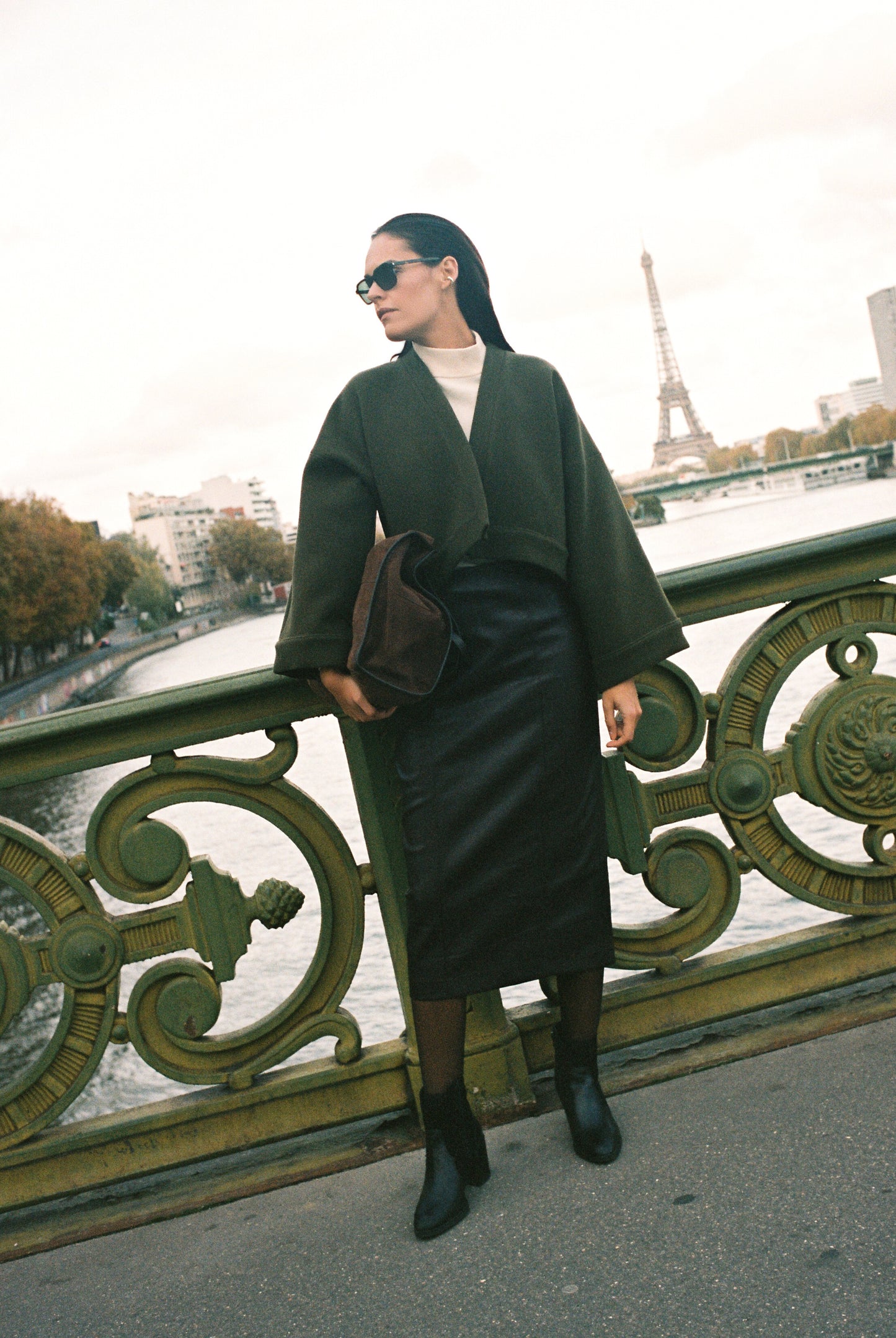 Femme portant le kimono court JOUFFROY Atelier Sarita en laine et cachemire kaki, coupe crop, avec finitions minimalistes et poches intérieures. La silhouette est photographiée sur un pont parisien avec vue sur la Tour Eiffel, mettant en valeur un kimono artisanal confectionné à la main à Paris, pensé pour l’hiver.