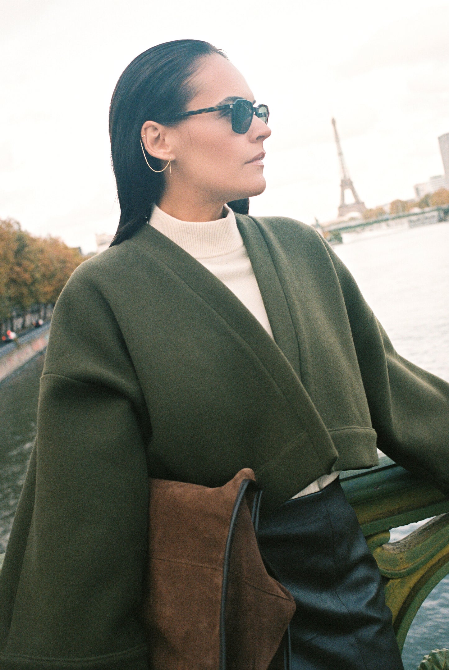 Femme portant le kimono court JOUFFROY Atelier Sarita en laine et cachemire kaki, coupe crop, avec finitions minimalistes et poches intérieures. La silhouette est photographiée sur un pont parisien avec vue sur la Tour Eiffel, mettant en valeur un kimono artisanal confectionné à la main à Paris, pensé pour l’hiver.