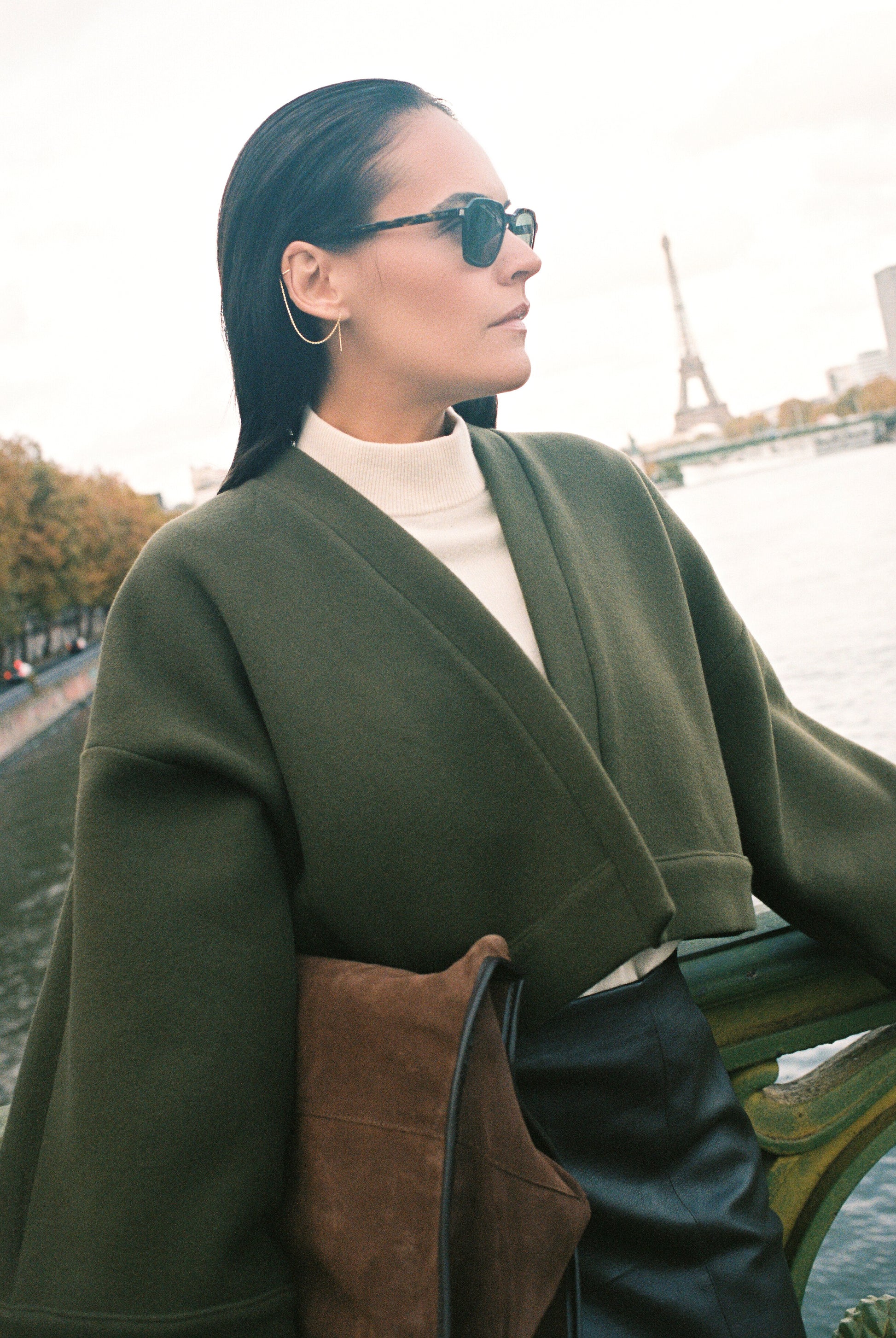 Femme portant le kimono court JOUFFROY Atelier Sarita en laine et cachemire kaki, coupe crop, avec finitions minimalistes et poches intérieures. La silhouette est photographiée sur un pont parisien avec vue sur la Tour Eiffel, mettant en valeur un kimono artisanal confectionné à la main à Paris, pensé pour l’hiver.