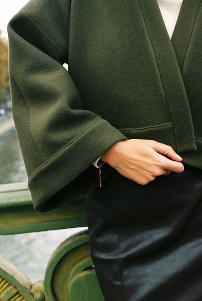 Femme portant le kimono court JOUFFROY Atelier Sarita en laine et cachemire kaki, coupe crop, avec finitions minimalistes et poches intérieures. La silhouette est photographiée sur un pont parisien avec vue sur la Tour Eiffel, mettant en valeur un kimono artisanal confectionné à la main à Paris, pensé pour l’hiver.