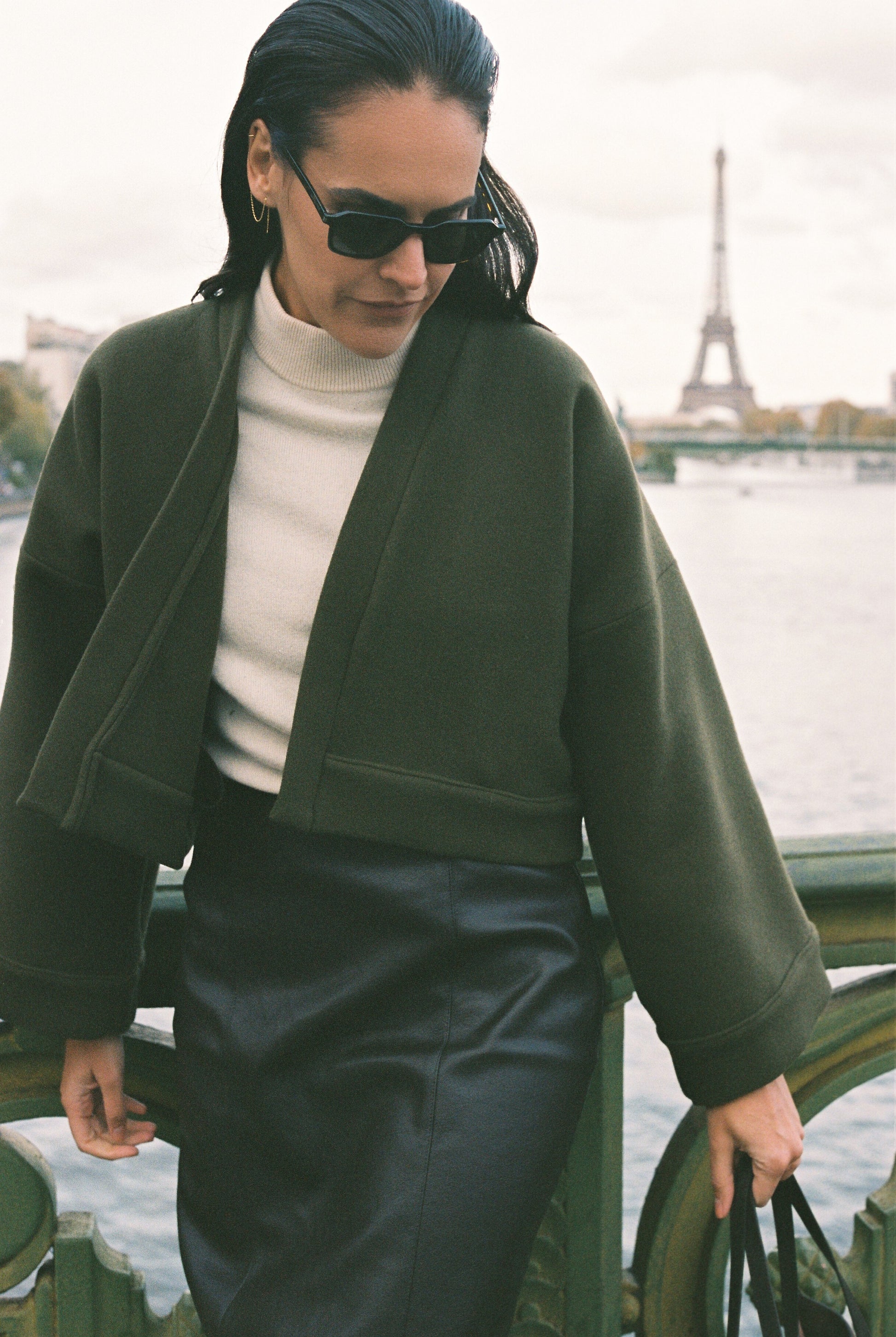 Femme portant le kimono court JOUFFROY Atelier Sarita en laine et cachemire kaki, coupe crop, avec finitions minimalistes et poches intérieures. La silhouette est photographiée sur un pont parisien avec vue sur la Tour Eiffel, mettant en valeur un kimono artisanal confectionné à la main à Paris, pensé pour l’hiver.