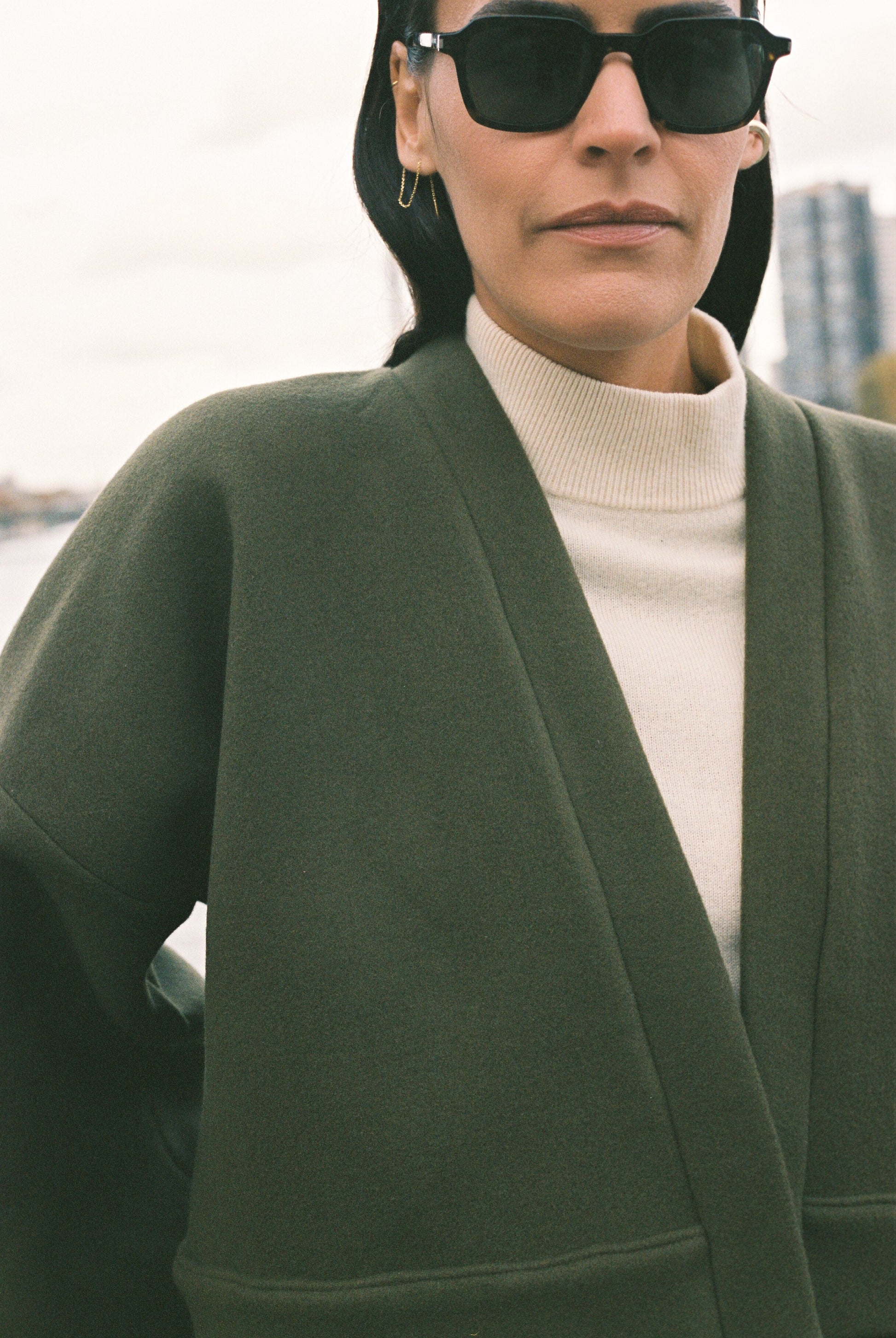 Femme portant le kimono court JOUFFROY Atelier Sarita en laine et cachemire kaki, coupe crop, avec finitions minimalistes et poches intérieures. La silhouette est photographiée sur un pont parisien avec vue sur la Tour Eiffel, mettant en valeur un kimono artisanal confectionné à la main à Paris, pensé pour l’hiver.