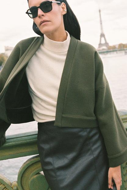 Femme portant le kimono court JOUFFROY Atelier Sarita en laine et cachemire kaki, coupe crop, avec finitions minimalistes et poches intérieures. La silhouette est photographiée sur un pont parisien avec vue sur la Tour Eiffel, mettant en valeur un kimono artisanal confectionné à la main à Paris, pensé pour l’hiver.