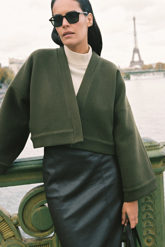 Femme portant le kimono court JOUFFROY Atelier Sarita en laine et cachemire kaki, coupe crop, avec finitions minimalistes et poches intérieures. La silhouette est photographiée sur un pont parisien avec vue sur la Tour Eiffel, mettant en valeur un kimono artisanal confectionné à la main à Paris, pensé pour l’hiver.