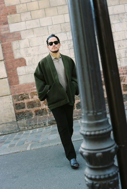 Homme portant le kimono RICHELIEU en laine et cachemire kaki d’Atelier Sarita, coupe veste avec grandes poches plaquées. Kimono structuré, fabriqué à la main à Paris dans un lainage italien issu de stocks dormants de maison de luxe. Modèle chaud, élégant et intemporel, photographié ans un style urbain parisien.
