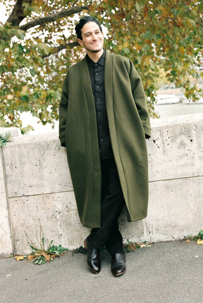 Homme portant le kimono long VIVIENNE en laine et cachemire kaki d’Atelier Sarita. Kimono oversize et structuré, fabriqué à la main à Paris dans un lainage italien issu de stocks dormants de maison de luxe. Pièce enveloppante et élégante, photographiée en extérieur avec la tour Eiffel en arrière-plan, mettant en valeur la douceur, la tenue et la chaleur du tissu.