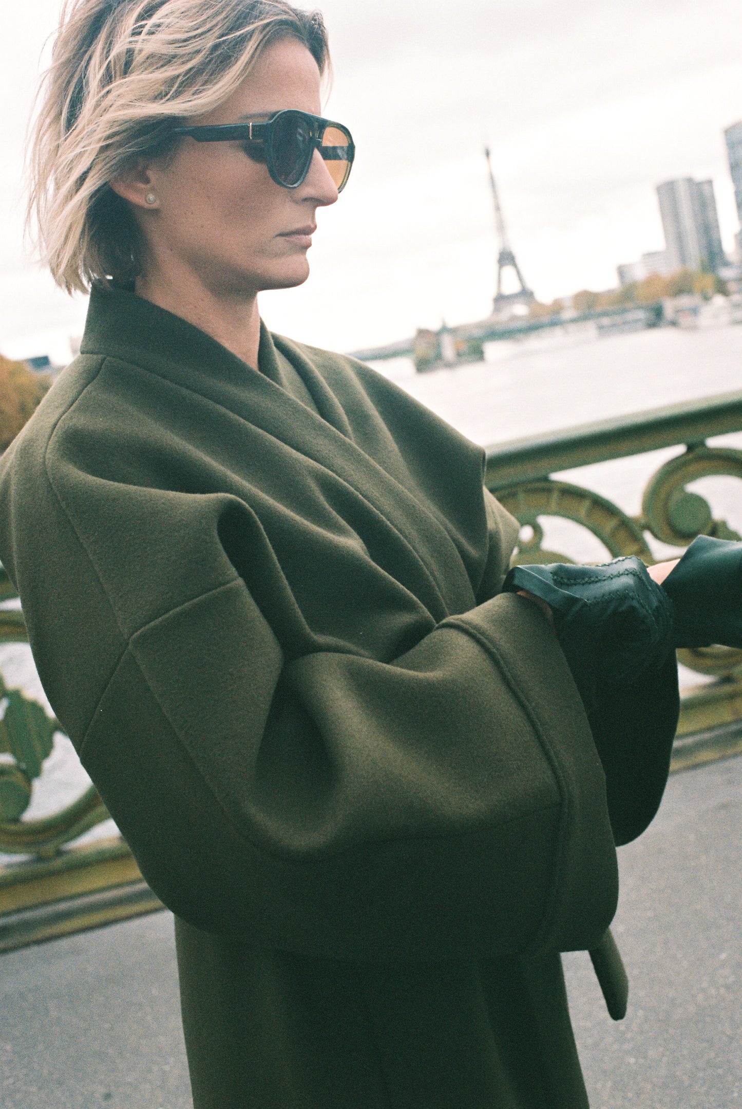 Femme portant le kimono long VIVIENNE en laine et cachemire kaki d’Atelier Sarita. Kimono oversize et structuré, fabriqué à la main à Paris dans un lainage italien issu de stocks dormants de maison de luxe. Pièce enveloppante et élégante, photographiée en extérieur avec la tour Eiffel en arrière-plan, mettant en valeur la douceur, la tenue et la chaleur du tissu.