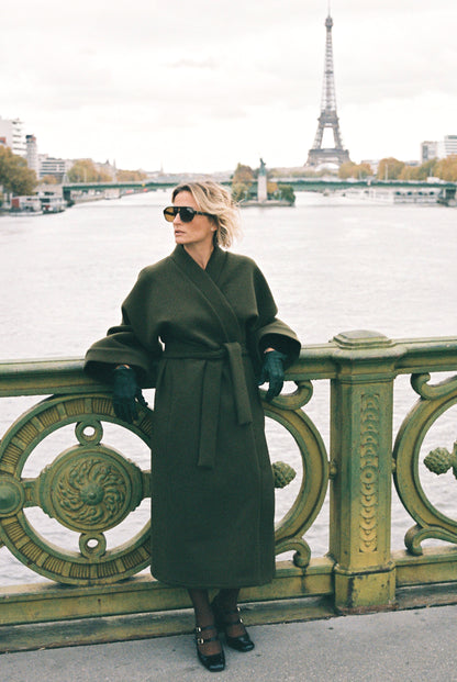 Femme portant le kimono long VIVIENNE en laine et cachemire kaki d’Atelier Sarita. Kimono oversize et structuré, fabriqué à la main à Paris dans un lainage italien issu de stocks dormants de maison de luxe. Pièce enveloppante et élégante, photographiée en extérieur avec la tour Eiffel en arrière-plan, mettant en valeur la douceur, la tenue et la chaleur du tissu.
