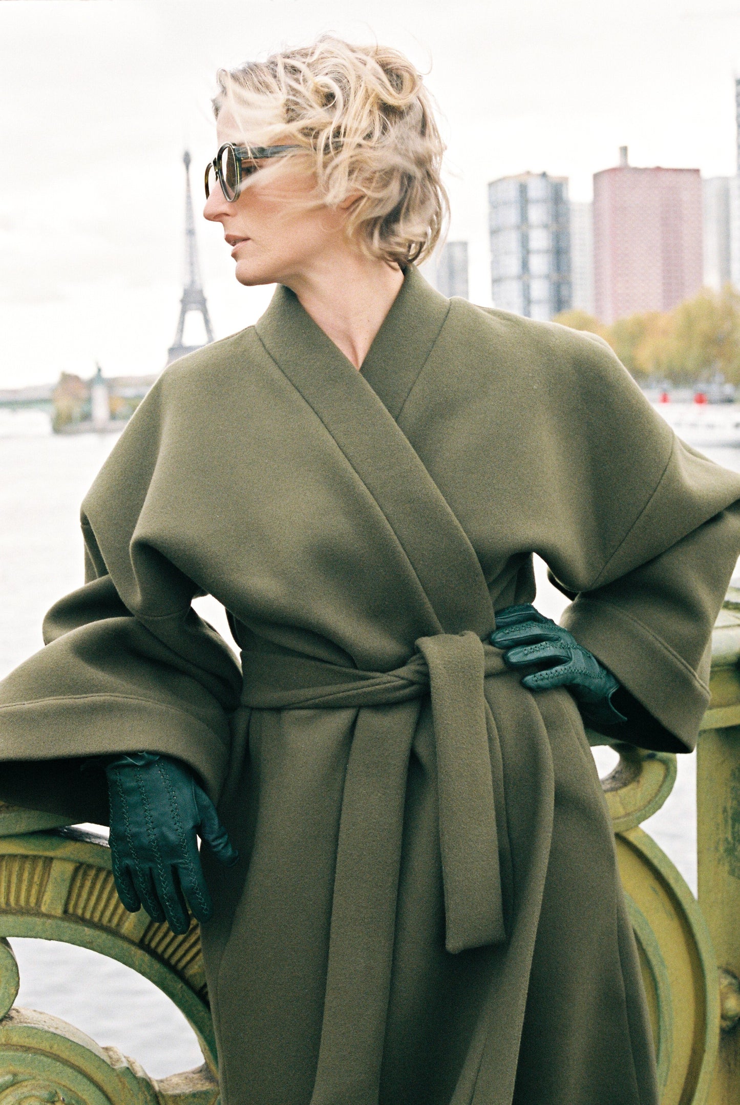 Femme portant le kimono long VIVIENNE en laine et cachemire kaki d’Atelier Sarita. Kimono oversize et structuré, fabriqué à la main à Paris dans un lainage italien issu de stocks dormants de maison de luxe. Pièce enveloppante et élégante, photographiée en extérieur avec la tour Eiffel en arrière-plan, mettant en valeur la douceur, la tenue et la chaleur du tissu.