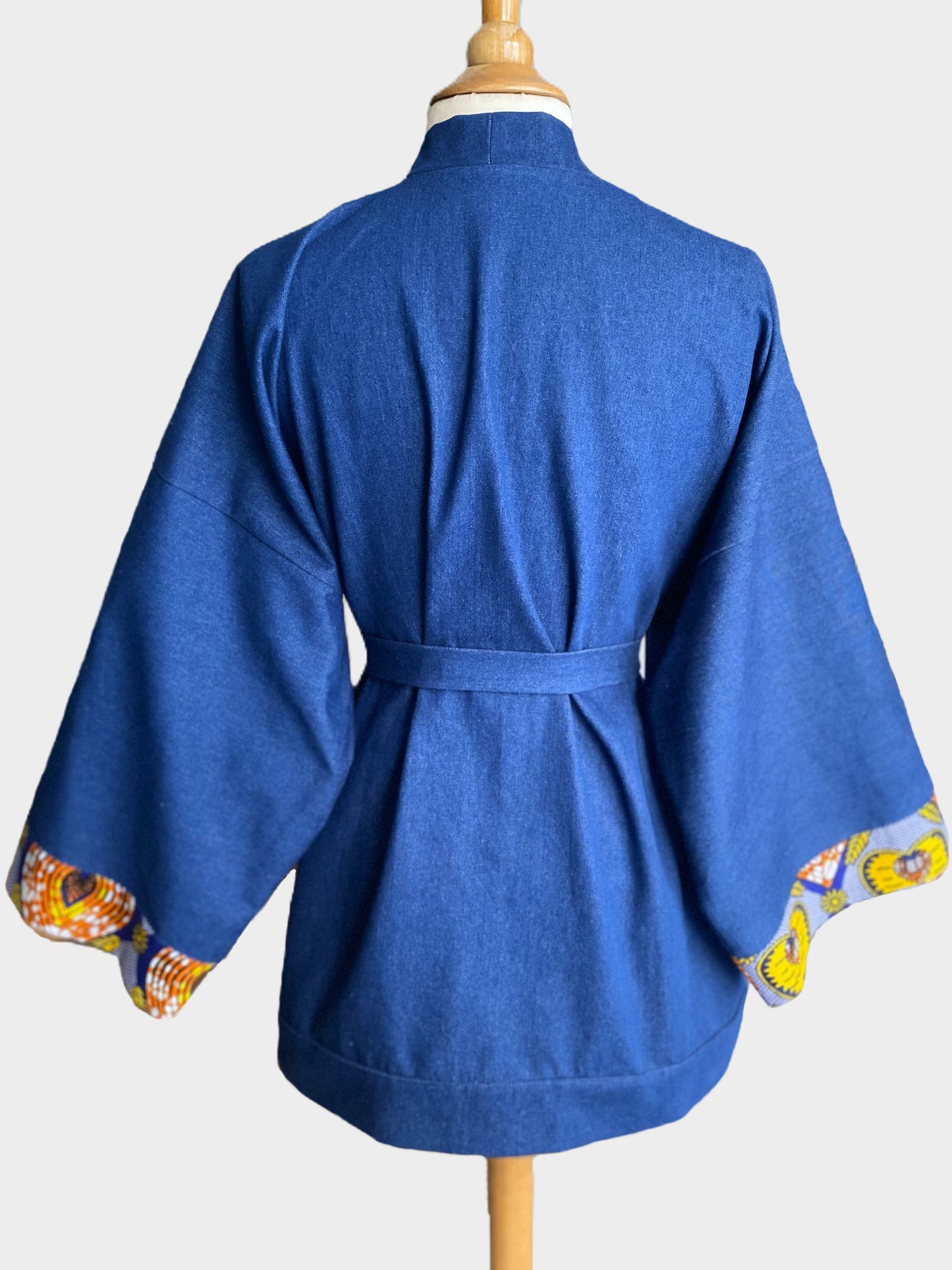 Kimono court en denim bleu avec finitions en tissu wax coloré sur les poignets et les poches, coupe ample et fluide.