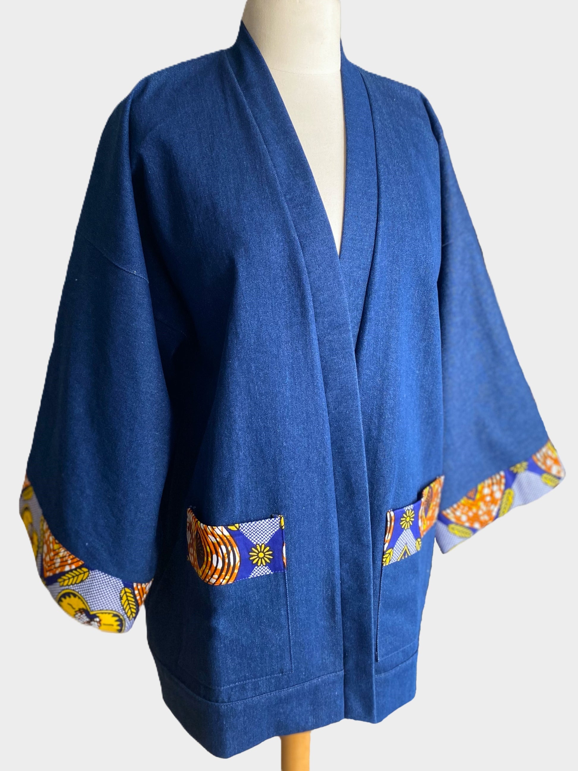 Kimono court en denim bleu avec finitions en tissu wax coloré sur les poignets et les poches, coupe ample et fluide.