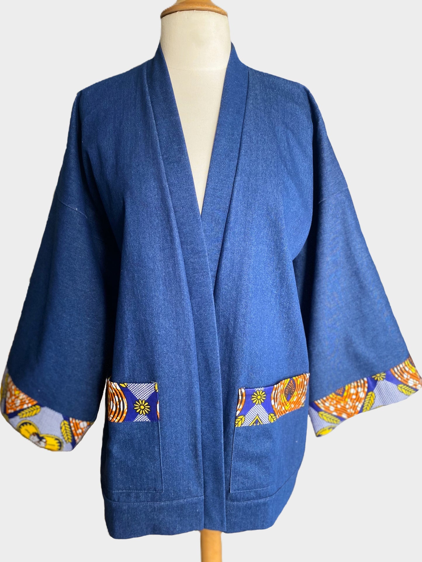 Kimono court en denim bleu avec finitions en tissu wax coloré sur les poignets et les poches, coupe ample et fluide.