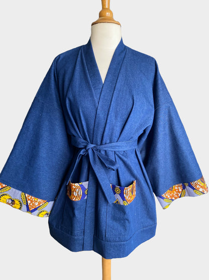 Kimono court en denim bleu avec finitions en tissu wax coloré sur les poignets et les poches, coupe ample et fluide.