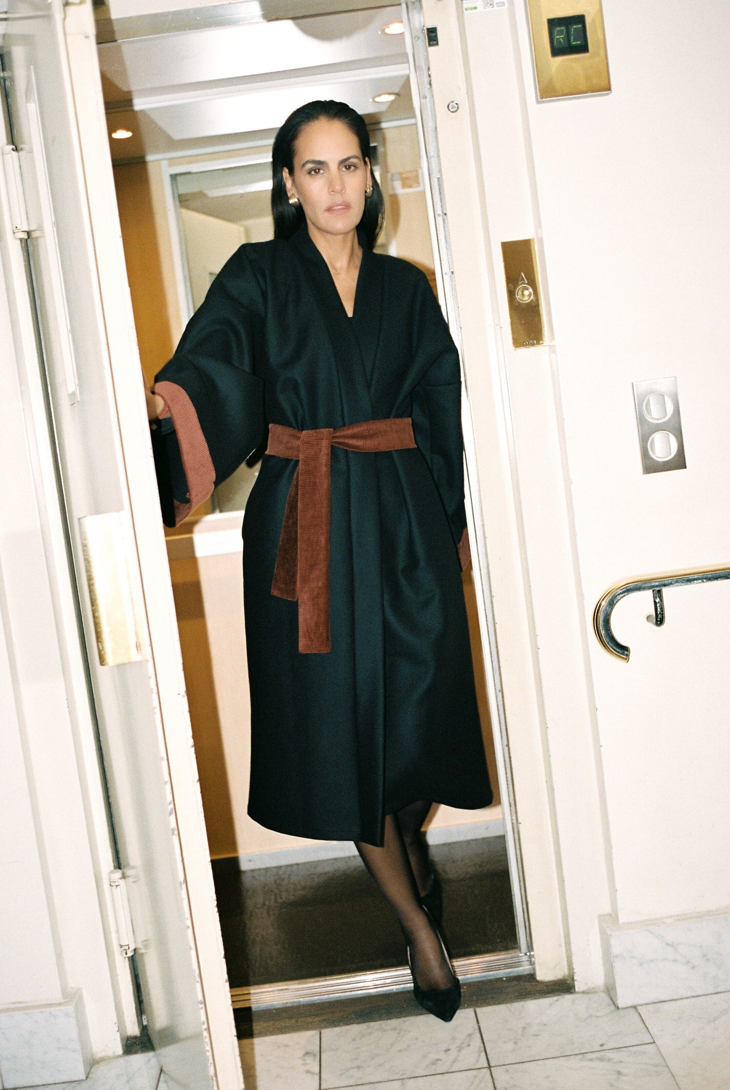 Femme portant le kimono long COLBERT Atelier Sarita en laine caviar noire avec finitions en velours coton couleur oud. Modèle ample et architectural, confectionné à la main à Paris, pensé comme un manteau kimono hivernal unisexe.