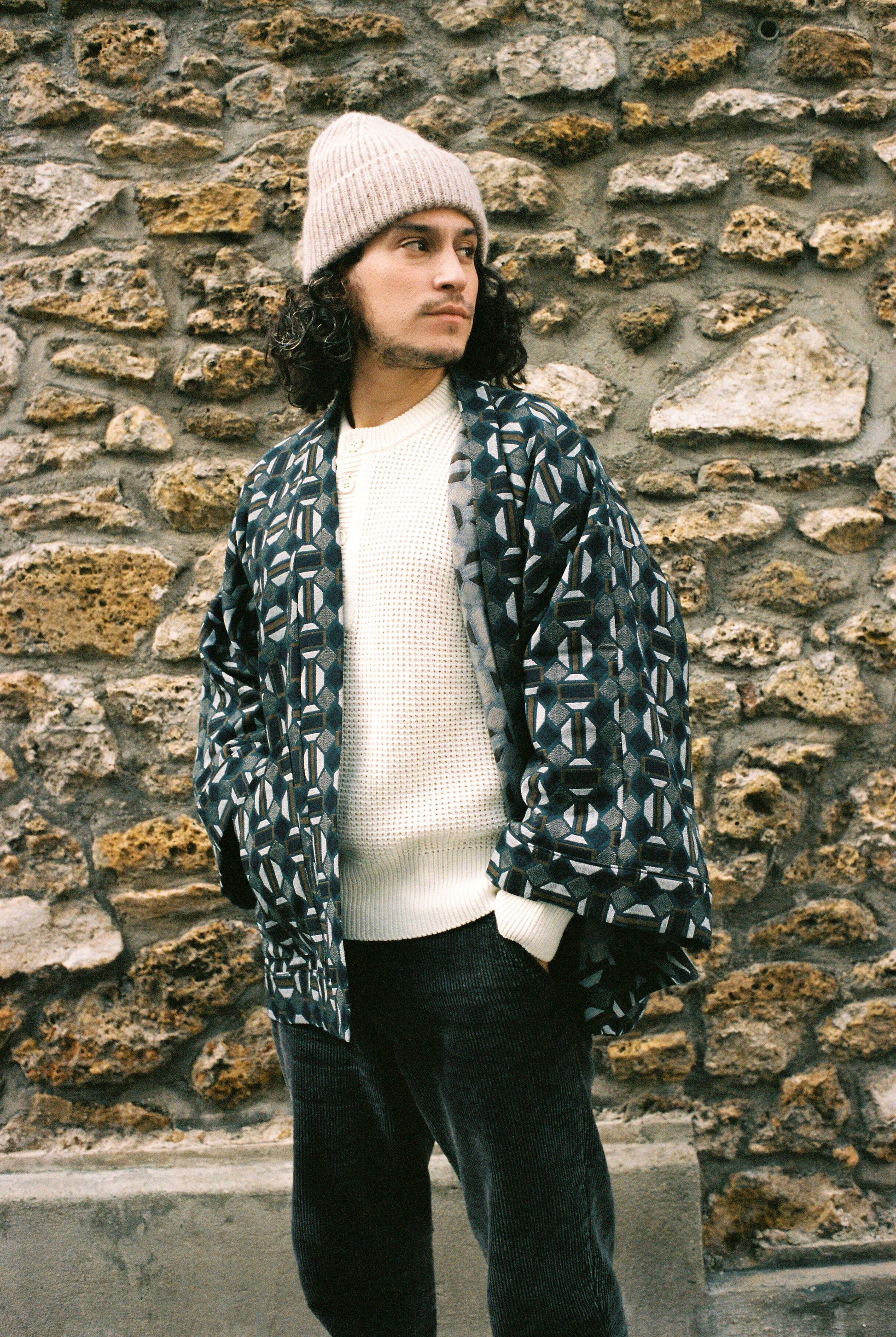 Homme portant le kimono VERDEAU Atelier Sarita, veste en jacquard géométrique bleu, gris et kaki, avec grandes poches plaquées. Kimono fabriqué à la main à Paris dans un jacquard issu d’une maison textile française historique. Photographie en extérieur devant un mur en pierre, mettant en valeur la texture dense du jacquard et la coupe ample et structurée du modèle.
