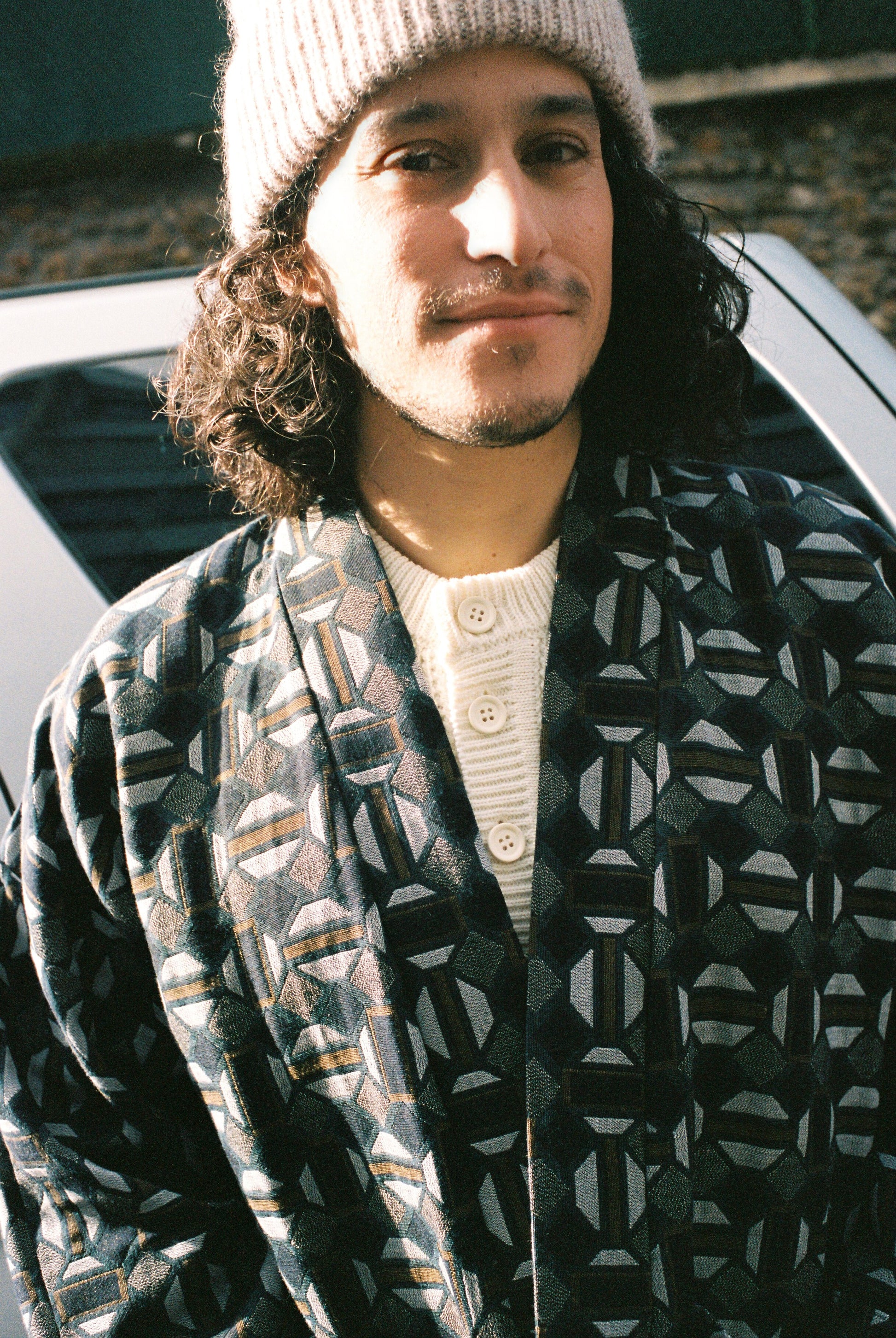 Homme portant le kimono VERDEAU Atelier Sarita, veste en jacquard géométrique bleu, gris et kaki, avec grandes poches plaquées. Kimono fabriqué à la main à Paris dans un jacquard issu d’une maison textile française historique. Photographie en extérieur devant un mur en pierre, mettant en valeur la texture dense du jacquard et la coupe ample et structurée du modèle.