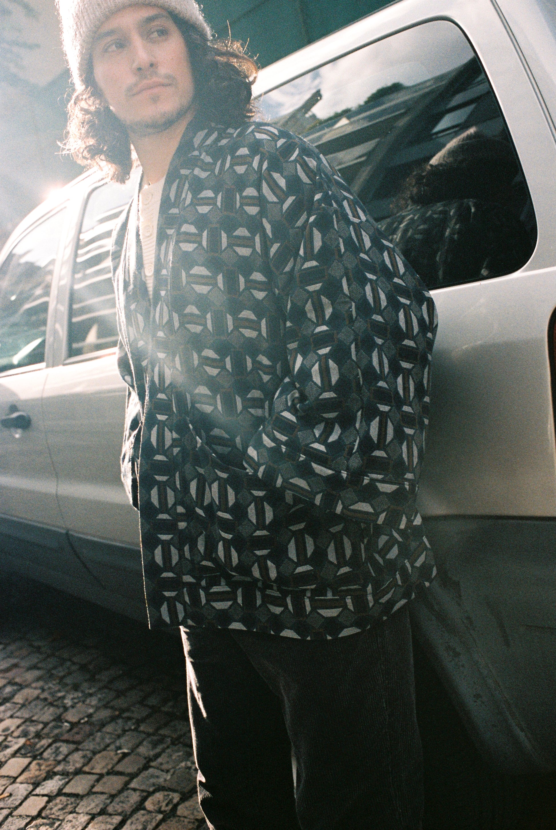 Homme portant le kimono VERDEAU Atelier Sarita, veste en jacquard géométrique bleu, gris et kaki, avec grandes poches plaquées. Kimono fabriqué à la main à Paris dans un jacquard issu d’une maison textile française historique. Photographie en extérieur devant un mur en pierre, mettant en valeur la texture dense du jacquard et la coupe ample et structurée du modèle.