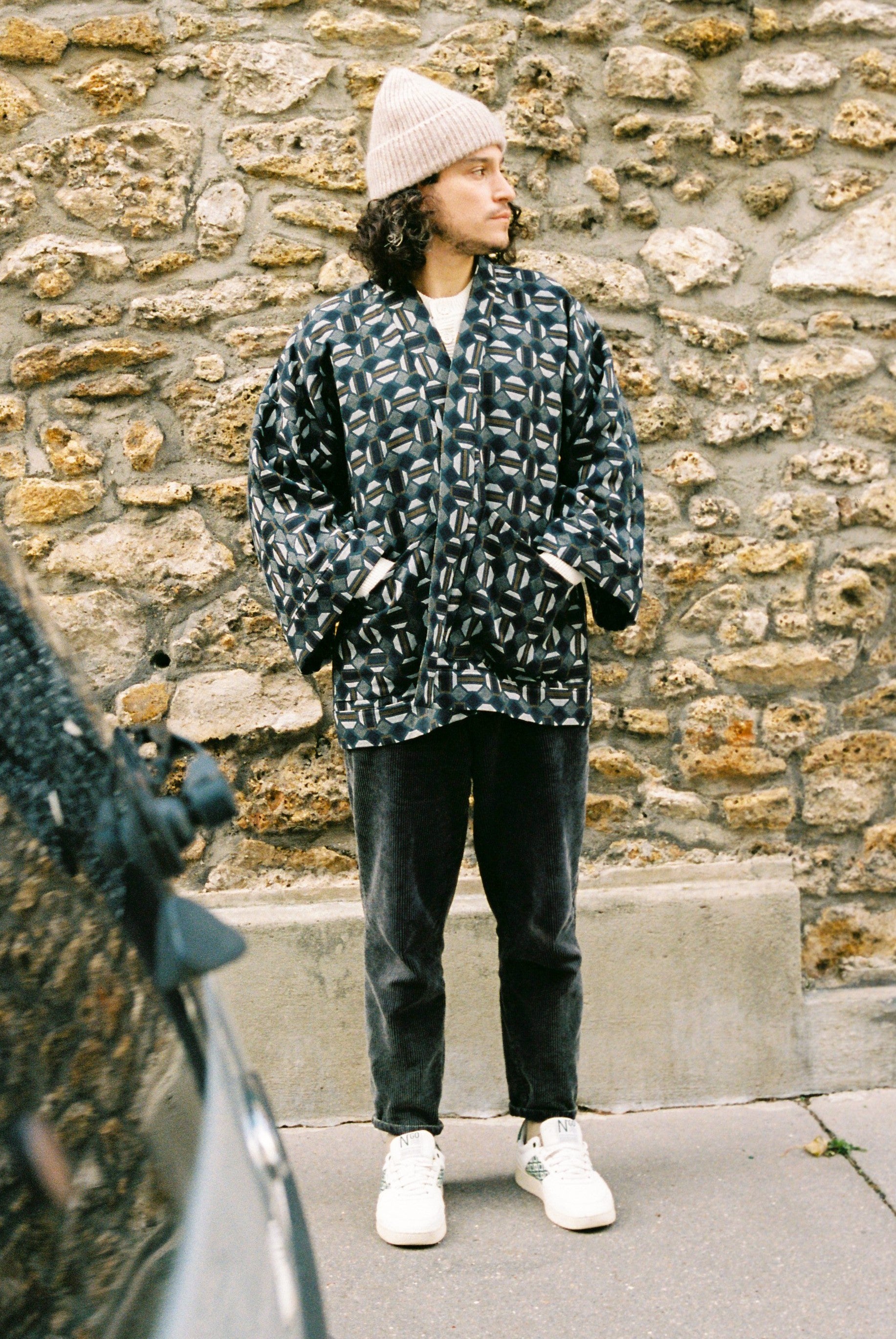 Homme portant le kimono VERDEAU Atelier Sarita, veste en jacquard géométrique bleu, gris et kaki, avec grandes poches plaquées. Kimono fabriqué à la main à Paris dans un jacquard issu d’une maison textile française historique. Photographie en extérieur devant un mur en pierre, mettant en valeur la texture dense du jacquard et la coupe ample et structurée du modèle.