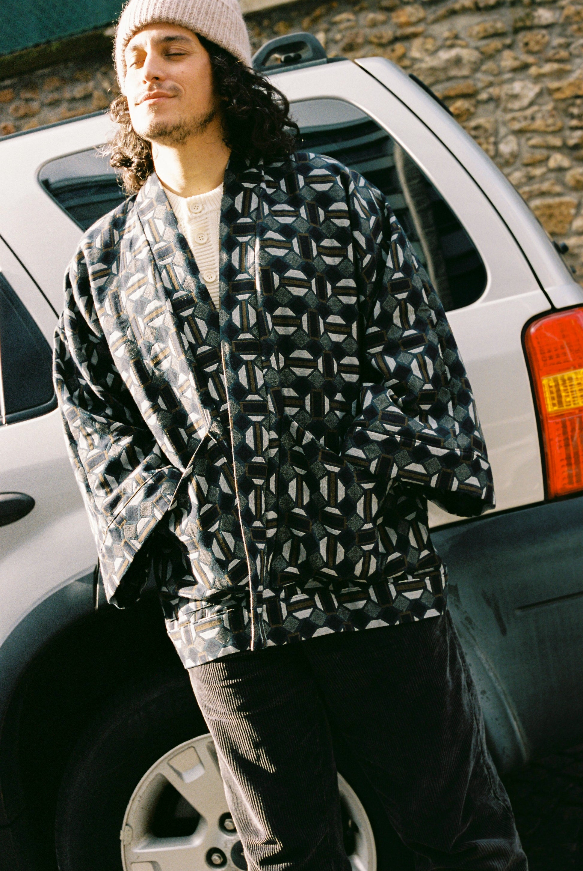 Homme portant le kimono VERDEAU Atelier Sarita, veste en jacquard géométrique bleu, gris et kaki, avec grandes poches plaquées. Kimono fabriqué à la main à Paris dans un jacquard issu d’une maison textile française historique. Photographie en extérieur devant un mur en pierre, mettant en valeur la texture dense du jacquard et la coupe ample et structurée du modèle.