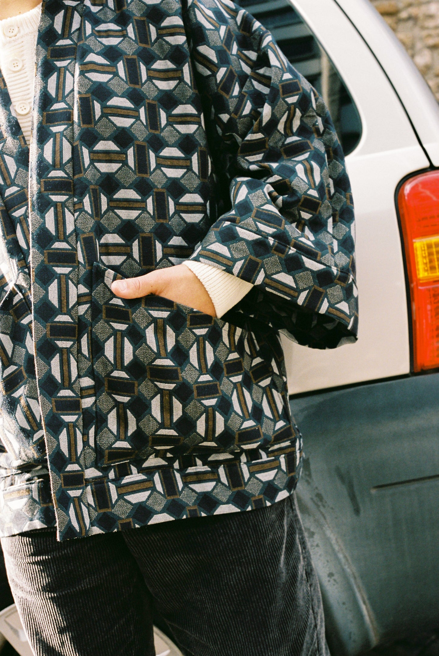 Homme portant le kimono VERDEAU Atelier Sarita, veste en jacquard géométrique bleu, gris et kaki, avec grandes poches plaquées. Kimono fabriqué à la main à Paris dans un jacquard issu d’une maison textile française historique. Photographie en extérieur devant un mur en pierre, mettant en valeur la texture dense du jacquard et la coupe ample et structurée du modèle.