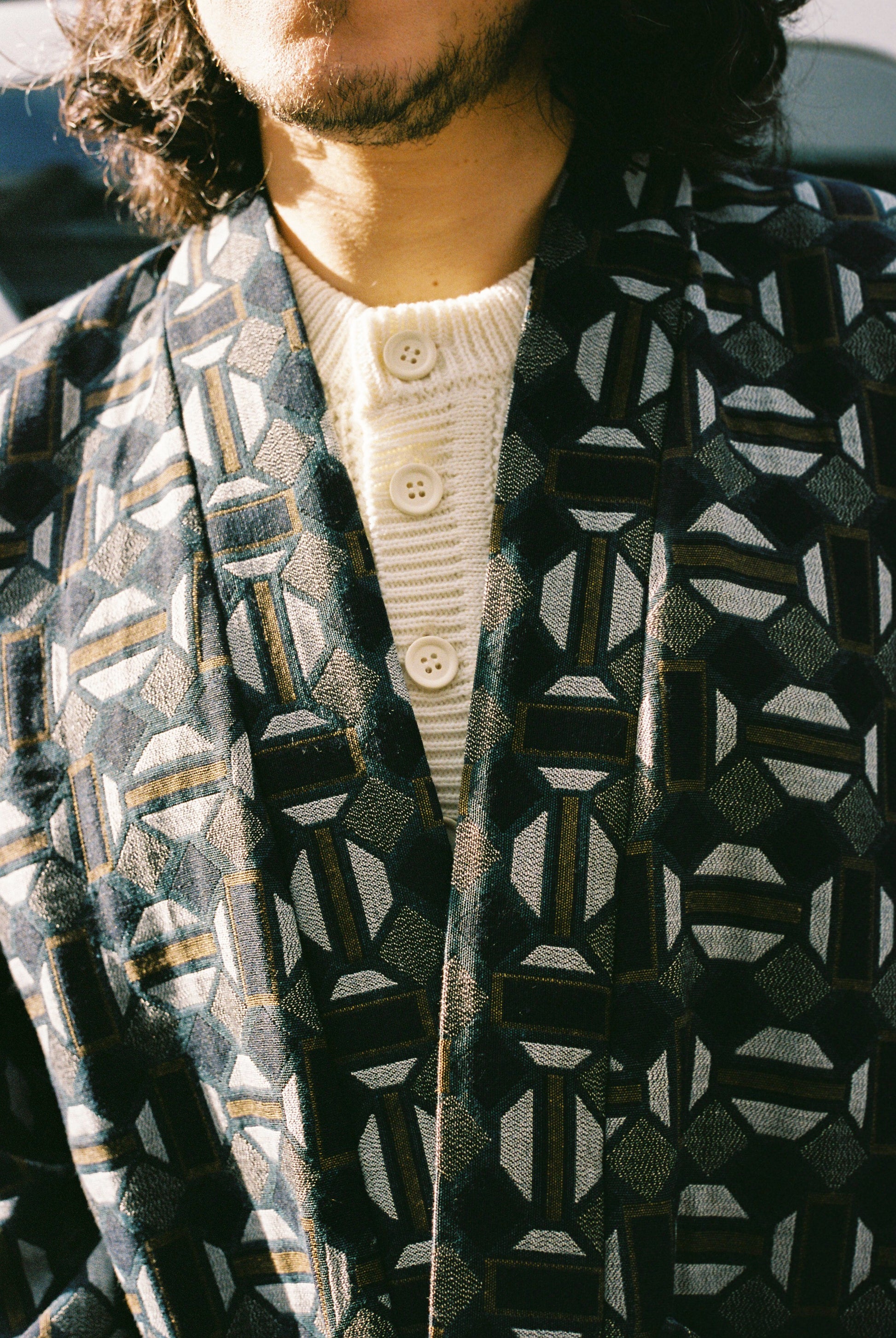 Homme portant le kimono VERDEAU Atelier Sarita, veste en jacquard géométrique bleu, gris et kaki, avec grandes poches plaquées. Kimono fabriqué à la main à Paris dans un jacquard issu d’une maison textile française historique. Photographie en extérieur devant un mur en pierre, mettant en valeur la texture dense du jacquard et la coupe ample et structurée du modèle.
