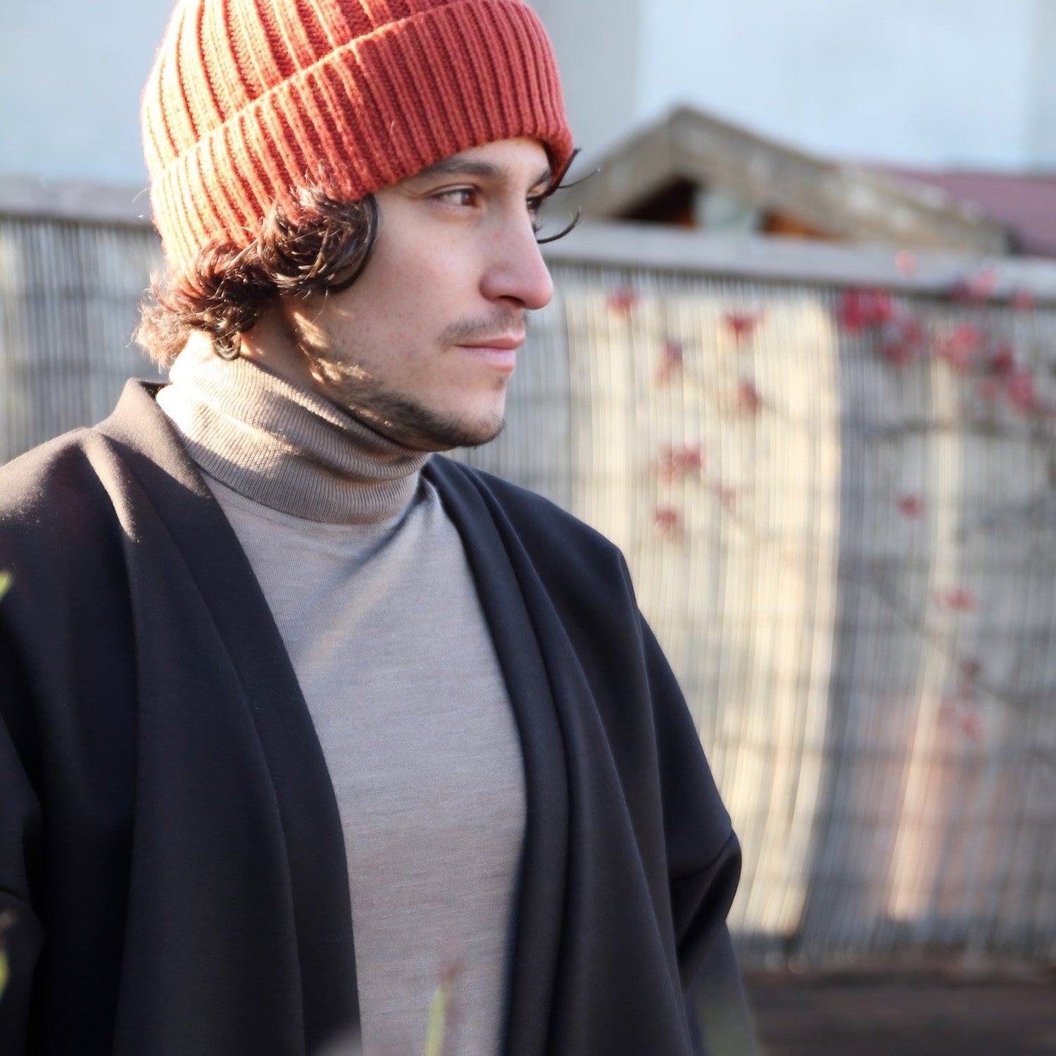 Homme portant un kimono noir Atelier Sarita, associé à un col roulé beige et un bonnet en laine orange. Une tenue élégante et intemporelle qui incarne l’authenticité et l’originalité des pièces uniques faites à la main, dans un cadre naturel et apaisant.